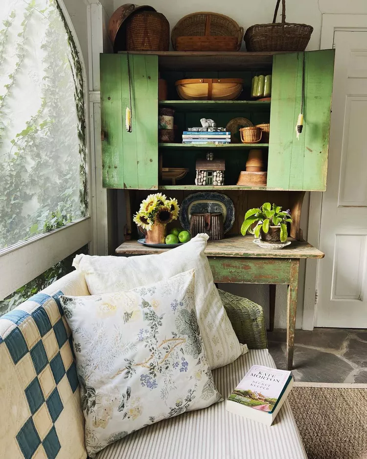 A clever storage setup showcasing small screened-in porch ideas decor, featuring a green distressed vintage cabinet filled with books and baskets for organization, placed next to a cozy daybed with floral accent pillows.