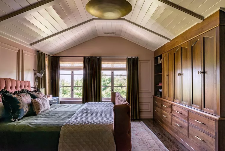 A cozy, luxury bedroom with a dusty pink tufted headboard, warm olive green velvet curtains, and massive custom built-in wooden wardrobes, crowned by a beautiful vaulted sloped ceiling lined with a crisp cream shiplap ceiling to show how horizontal wooden groove layouts add classic structural craftsmanship, movement, and coastal cottage charm.