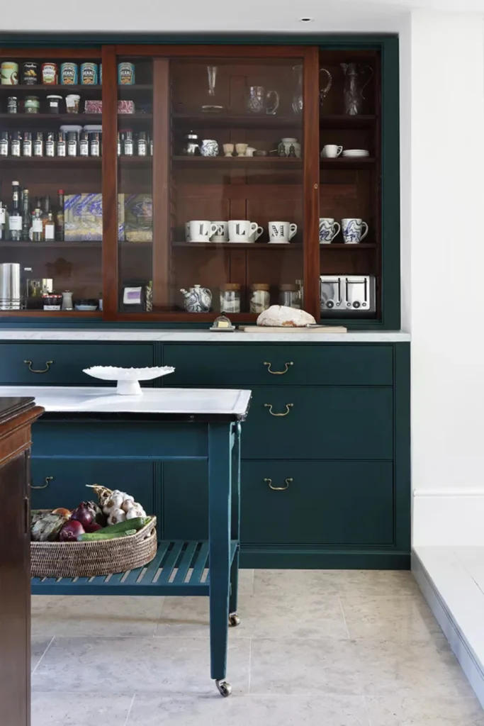 A rustic modern kitchen space showcasing creative navy blue accent wall ideas through deep navy-painted built-in lower cabinets with brass handles. The setup features a white marble countertop under a traditional wooden display cabinet with glass sliding doors, alongside a matching navy blue rolling prep cart holding a fresh vegetable basket on a light stone floor.