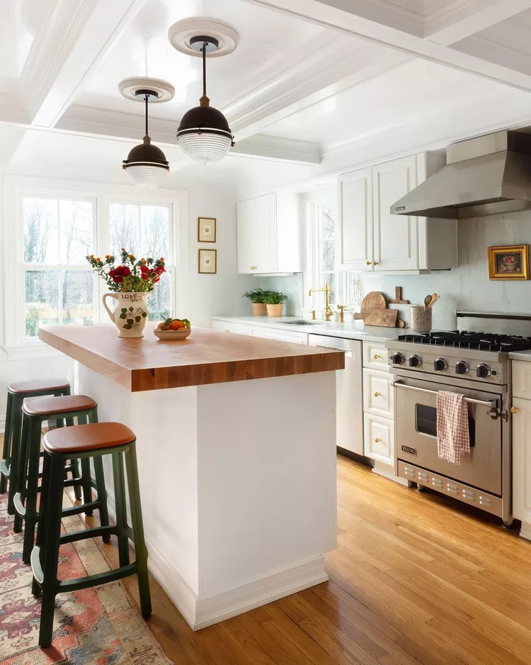 A bright, clean kitchen featuring a white kitchen island with a butcher block countertop and green bar stools, highlighted by a simple coffered ceiling painted in a uniform, crisp white gloss with recessed panels and matching pendant light fixtures to create structural depth and timeless architectural appeal.