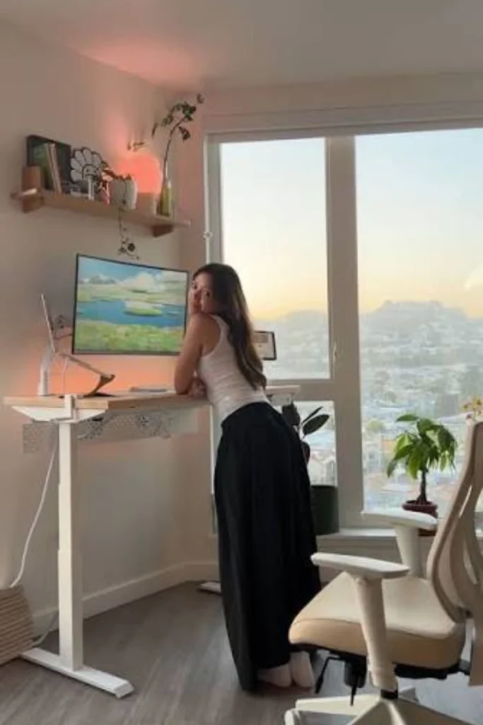 A woman working at a modern height-adjustable desk near a large window with city views, highlighting ergonomic home office ideas for women sit stand desk setups featuring a motorized white frame, light wood top, and a matching white ergonomic desk chair.