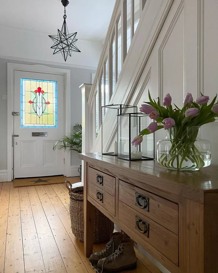A bright entryway hallway with a rustic wooden console table and purple tulips, leading toward a white front door featuring a beautifully detailed floral stained glass window pane that serves as a permanent architectural art piece.