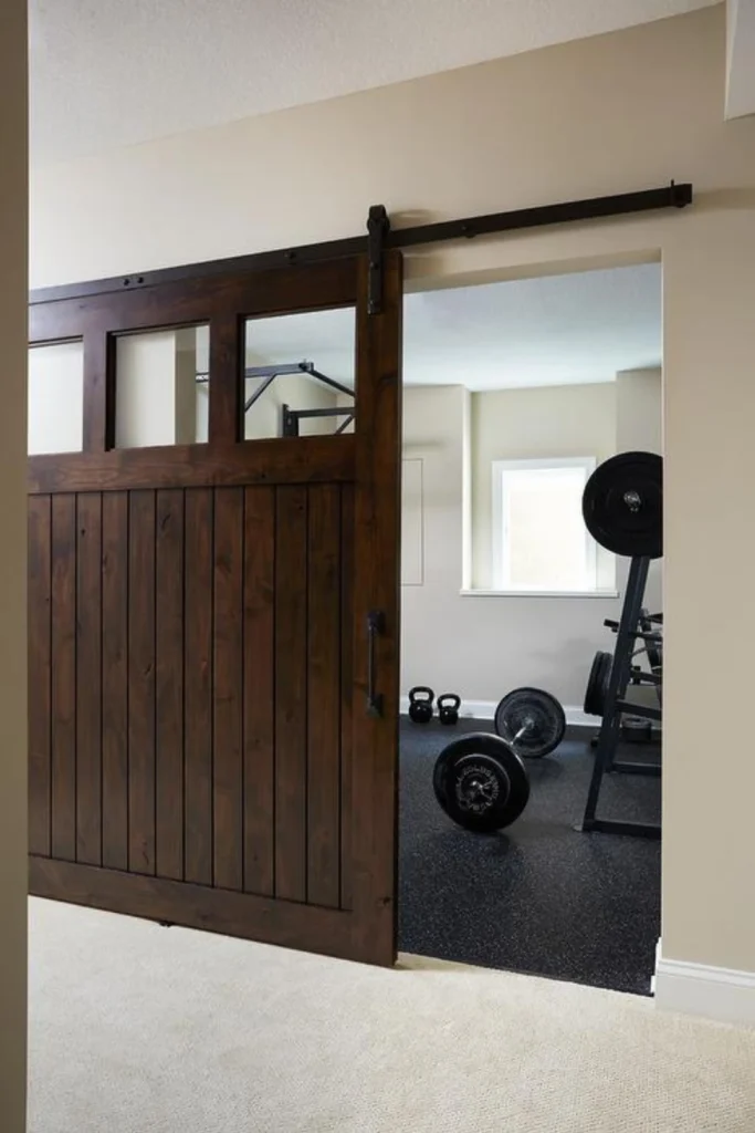 A rustic sliding barn door with dark wood paneling and glass windows partially slid open to reveal a home gym space with dark rubber mat flooring, kettlebells, a barbell, and a weight rack.