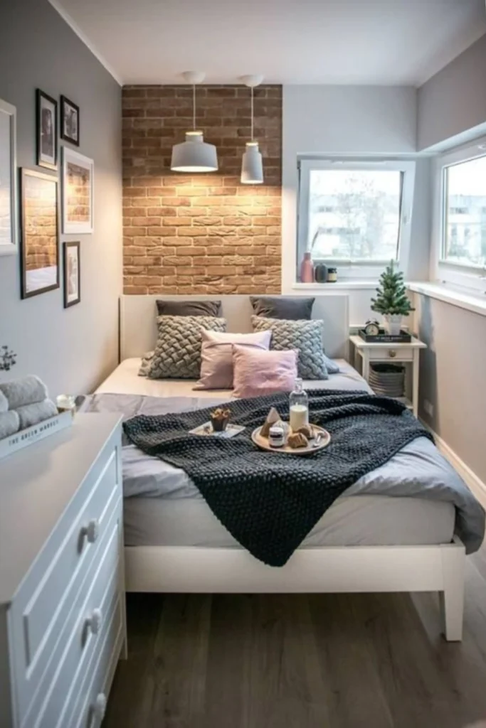 A small, cozy guest bedroom featuring a white bed frame with soft grey and pink pillows, an exposed brick accent wall with hanging pendant lights, and clean-lined inside-mount window shades, showing how Dressing small windows in tight spaces requires a clever balance of functionality, proportion, and visual lightness. Finding smart window treatment ideas for small guest bedrooms ensures your visitors feel welcomed in a charming, cozy space without making the room feel cramped or dark. In a smaller guest room, bulky drapes can quickly swallow up precious wall space. Instead, lean into clean-lined treatments like fitted cellular shades, woven bamboo blinds, or tailored Roman shades that fit neatly inside the window frame. Inside-mount shades: Keeps the walls clear and visual clutter to an absolute minimum. Match the wall color: Pick curtain panels in the same shade as your paint to create a seamless, room-expanding look. Vertical stripes: Choose subtle striped fabrics to draw the eye upward and create an illusion of height.