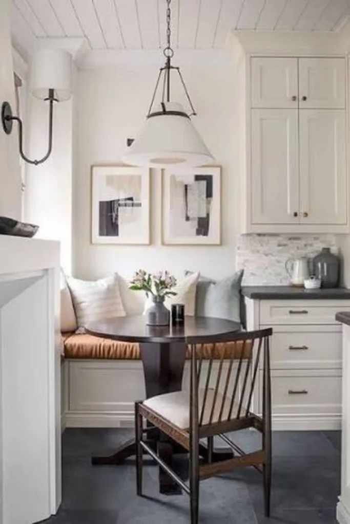 A compact kitchen corner featuring a cozy Space-Saving Breakfast Nook. The design highlights a small round dark wood pedestal table paired with a single wooden chair and a built-in white banquette bench with tan cushions, grey throw pillows, and two abstract framed art prints on the wall beneath a hanging cone-shaped pendant light.