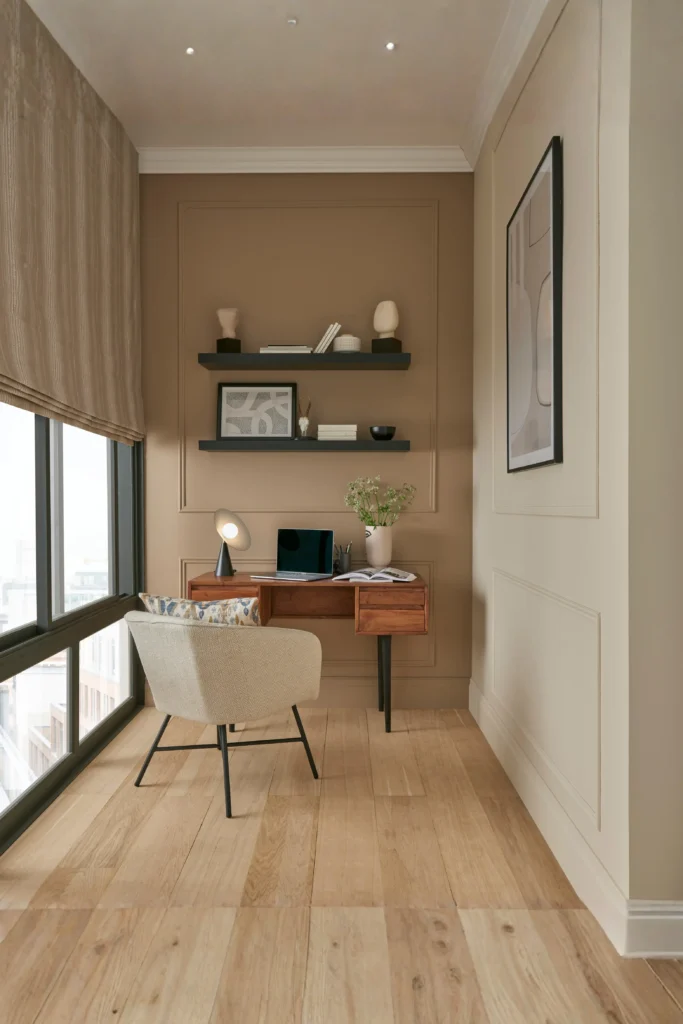 A cozy modern home office setup showcasing warm minimalist women home office ideas interior design, featuring a wooden desk placed perpendicular to a floor-to-ceiling window, a beige armchair, floating black shelves, and neutral warm wall paneling.