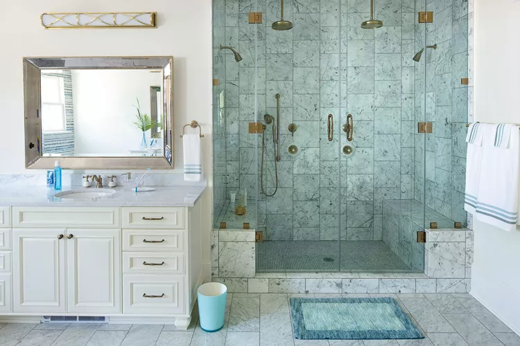 A luxury custom master bathroom layout highlighting why you should splurge on the shower enclosure. The space features a massive walk-in shower with seamless, frameless glass panels, clad entirely in beautiful gray-veined white marble tiles. The shower includes dual brass rainfall showerheads and a built-in marble bench, perfectly positioned next to a classic white vanity cabinet with a matching marble countertop and a wide metallic mirror.