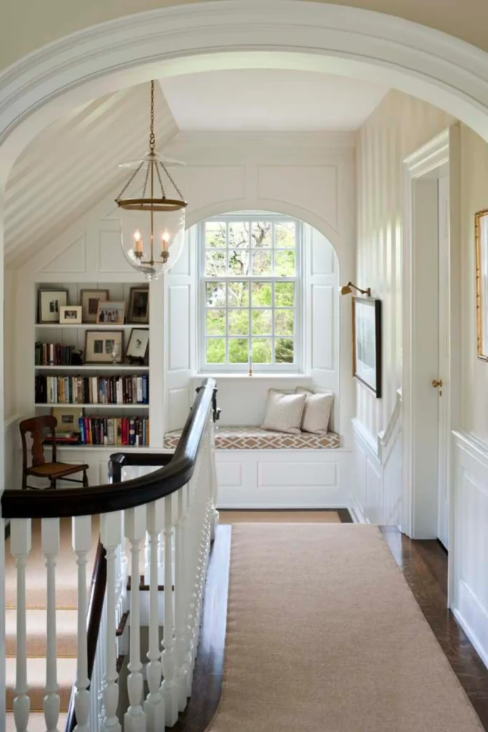 A bright home interior displaying elegant staircase landing ideas with window features, showcasing a custom white built-in bench seat under a large arched window. The cozy stairwell nook is styled with neutral throw pillows, built-in bookshelves packed with books, traditional white wall wainscoting, and a vintage brass hanging lantern light.