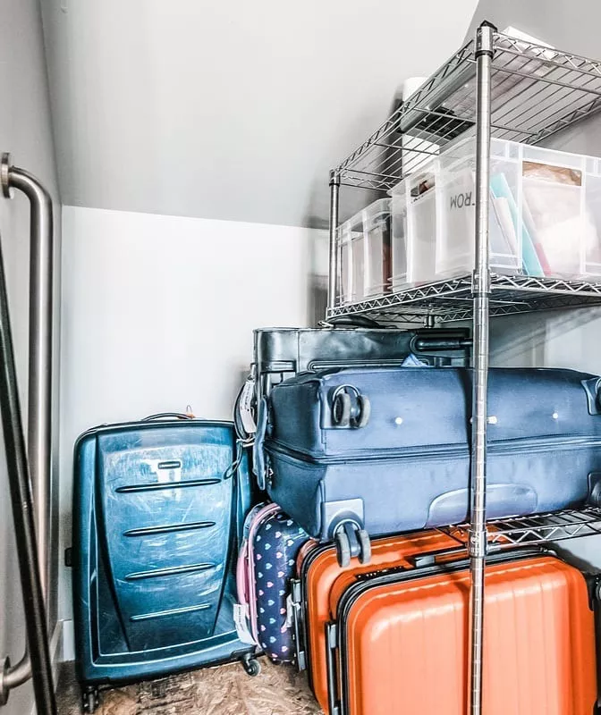 A close-up view of an attic eave storage space under a low angled ceiling, packed with large travel suitcases. An orange hardside suitcase and multiple dark blue and black rolling suitcases are stacked vertically alongside a silver wire utility rack holding clear plastic organizational bins, demonstrating how to use deep, hard-to-reach triangular spaces to store luggage out of the way.