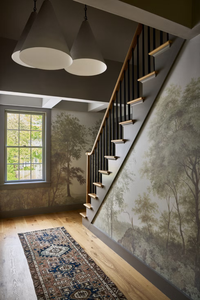 A gorgeous home entryway demonstrating the Textural Illusion: Bringing the Outdoors In concept. The walls along the hallway and under the wooden staircase feature a realistic, moody panoramic forest mural wallpaper depicting tall green trees. Light oak hardwood floors are complemented by a patterned bohemian runner rug, illuminated by large white cone pendant lights hanging from the ceiling.