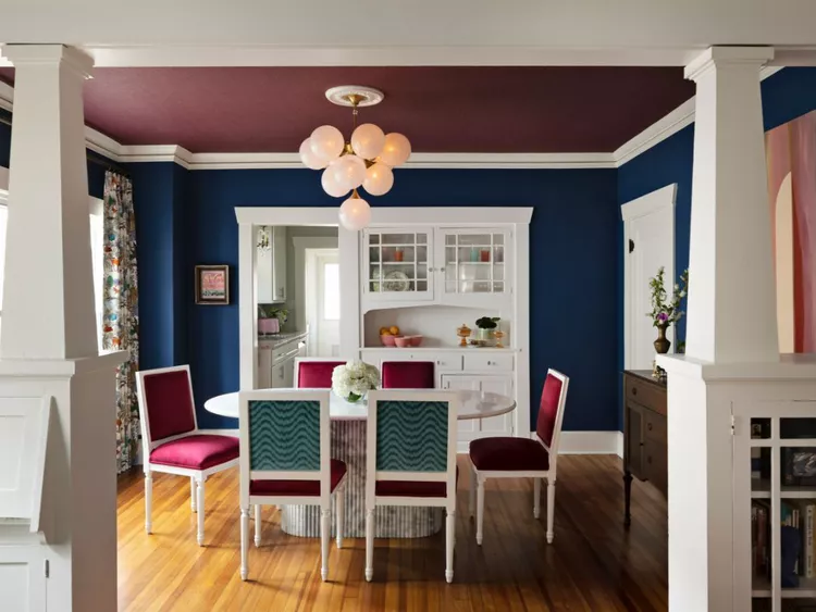 A sophisticated dining room with deep blue walls, a white built-in hutch, and dining chairs upholstered in rich magenta and patterned teal, styled with a solid plum-colored textural wallpapered ceiling that brings a soft, tactile dimension and subtle depth to the room without overwhelming the colorful design.