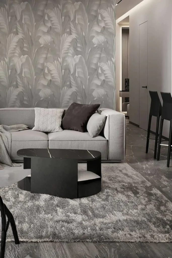 A sophisticated modern living room showcasing the latest gray living room wallpaper trends 2026. The space features a prominent accent wall covered in a textured gray banana leaf pattern wallpaper. A plush light gray sofa with patterned throw pillows is paired with a sleek black marble coffee table on a thick gray area rug, creating a quiet luxury aesthetic.