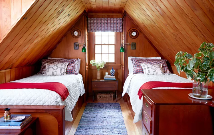 A cozy attic bedroom layout with twin beds displaying smart bedroom flooring ideas by choosing to try a runner rug instead of a full area rug. The space features light hardwood floors with a long, textured blue woven runner rug laid in the center walkway between the two beds. The room is designed with warm, matching slanted wood-paneled walls and ceilings, a small wooden nightstand under a central window, and vibrant red throws on each bed.