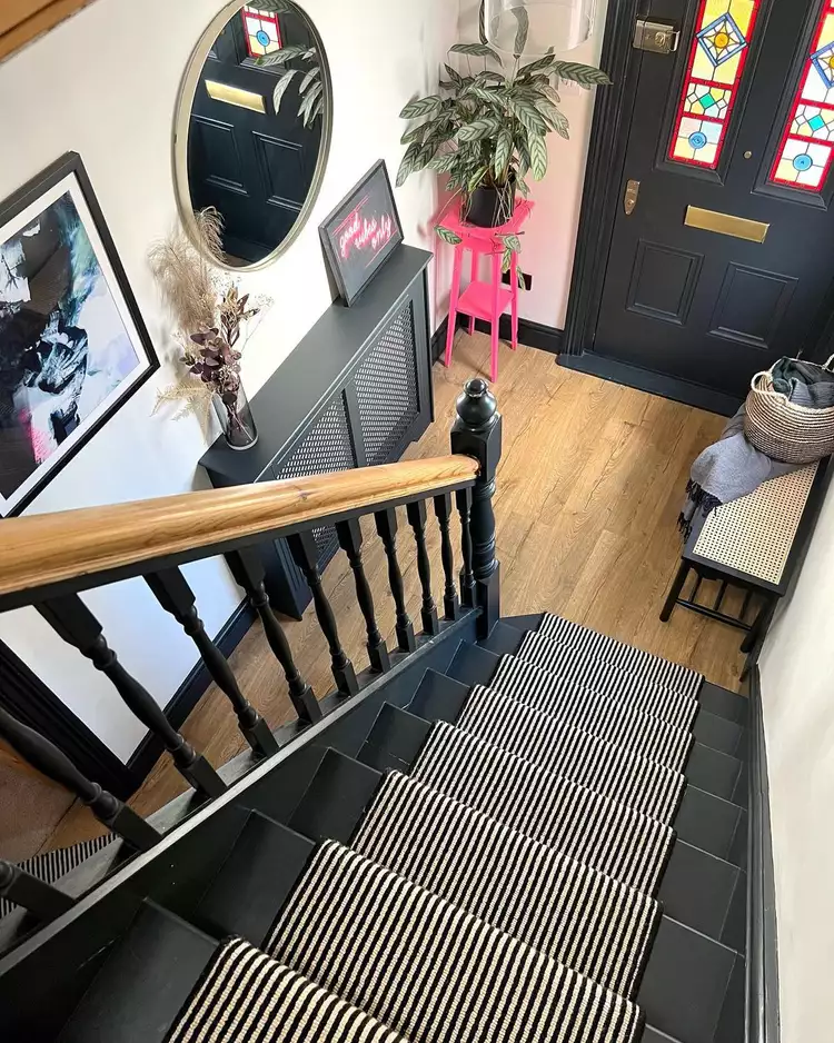 An overhead view of a dark wooden staircase looking down onto an entryway foyer, featuring a black front door with colorful geometric stained glass window panels that add vintage charm to the hallway layout.