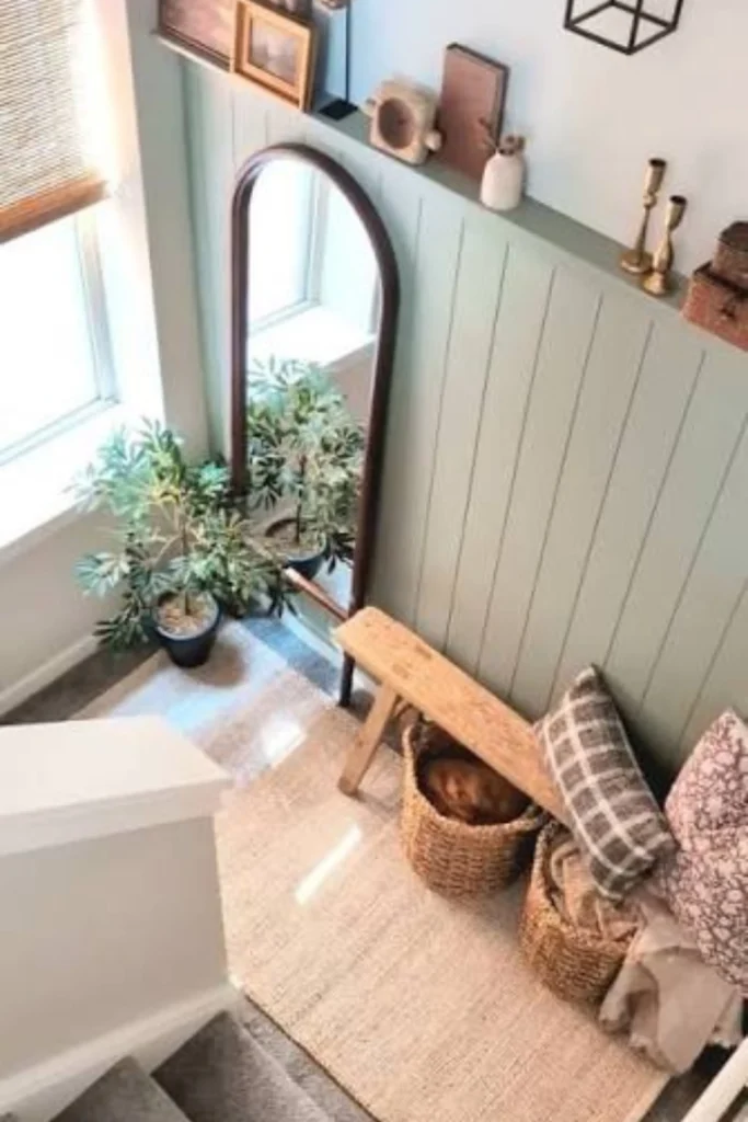 A bright stairwell corner showcasing creative staircase landing ideas with window elements. The space features a rustic light wood bench styled with plaid and floral throw pillows, resting on a natural jute rug. Next to a sunlit window with bamboo shades, a tall arched wood-framed floor mirror sits near a potted green plant, set against a sage green shiplap accent wall decorated with small framed art and brass candlesticks.