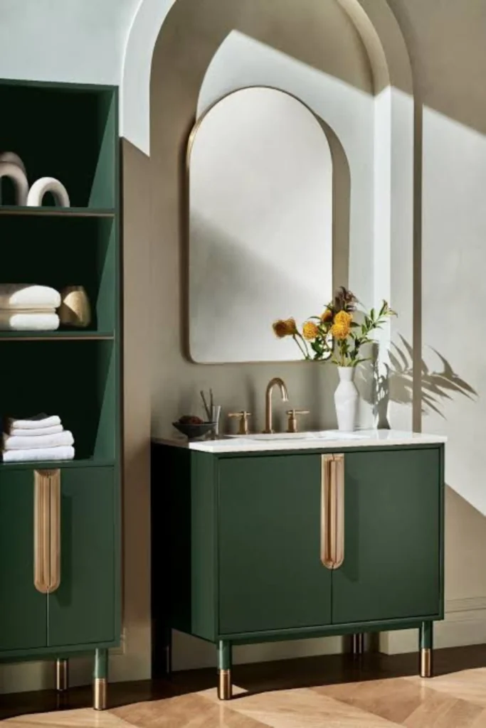 A sophisticated upscale vanity layout showing elegant bathroom remodel ideas modern designs. A sleek, emerald green vanity unit with modern long brass handle bars and a white marble countertop stands beautifully beneath a large arched wall mirror. The setup is nested against a clean beige wall with a striking architectural arch recess, right next to a matching green storage shelf unit on rich herringbone hardwood floors.