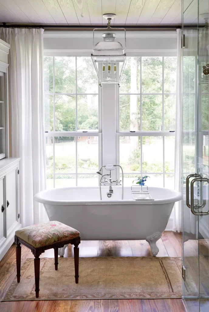 A bright master bathroom displaying how to use antique fixtures to add timeless character to a layout. The space features a freestanding white clawfoot soaking tub with polished vintage-style metal taps, positioned in front of large double windows with white curtains. An ornate upholstered antique stool rests on a neutral vintage rug over warm hardwood floors, under a hanging industrial lantern pendant light.