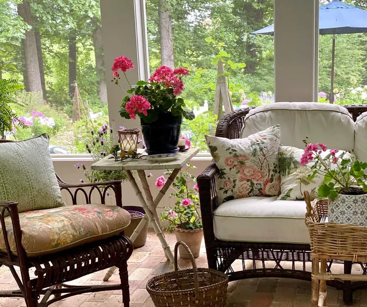 A cozy small screened-in porch featuring space-saving folding furniture, with a rustic white wooden folding table holding a pot of pink geraniums, placed between comfortable dark wicker chairs with floral accent pillows.