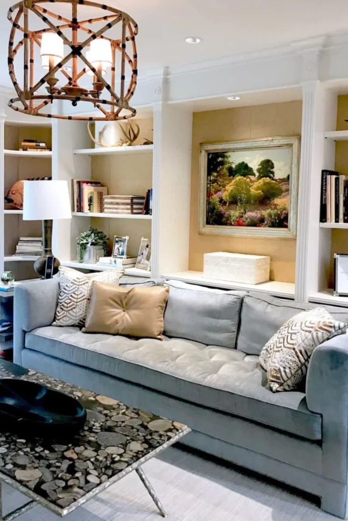 A cozy living room demonstrating small house decorating ideas living room modern layouts. A large plush light gray tufted sofa acts as the main focal point, styled with patterned throw pillows and a tan leather cushion. The background features built-in white bookshelves with an elegant oil painting, a modern floor lamp, and a unique bamboo pendant chandelier overhead.