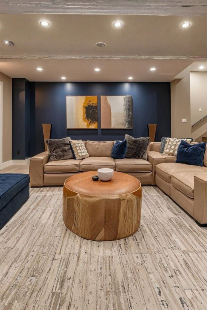 A cozy, modern finished basement showcasing brilliant navy blue accent wall ideas. The room features a large beige sectional sofa decorated with dark grey and navy velvet pillows, centered around a unique round wooden coffee table with a leather top. Two abstract paintings hang on the deep blue accent wall under warm recessed ceiling spotlights, all resting on a textured light grey floor.