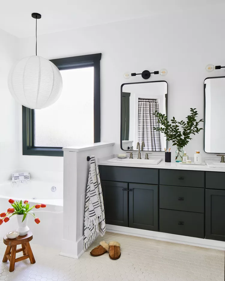 A modern monochrome bathroom layout displaying how to use stark contrast to create a high-impact aesthetic. The space features a dark charcoal black vanity with clean white countertops under twin black-framed rectangular mirrors. Crisp white walls and light hexagon floor tiles contrast sharply with a black window frame, while a large white paper globe pendant light hangs over a built-in white soaking tub.