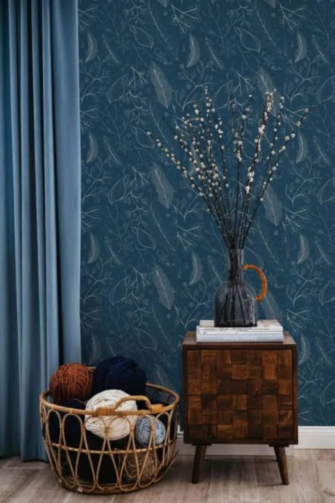A cozy room corner demonstrating elegant navy blue accent wall ideas with an organic patterned botanical wallpaper featuring a rich navy blue background and white line illustrations. Next to a light blue curtain, a textured dark wooden nightstand holds a tall glass vase with decorative pussy willow branches, complemented by a large wicker basket filled with colorful yarn balls on a light wood floor.