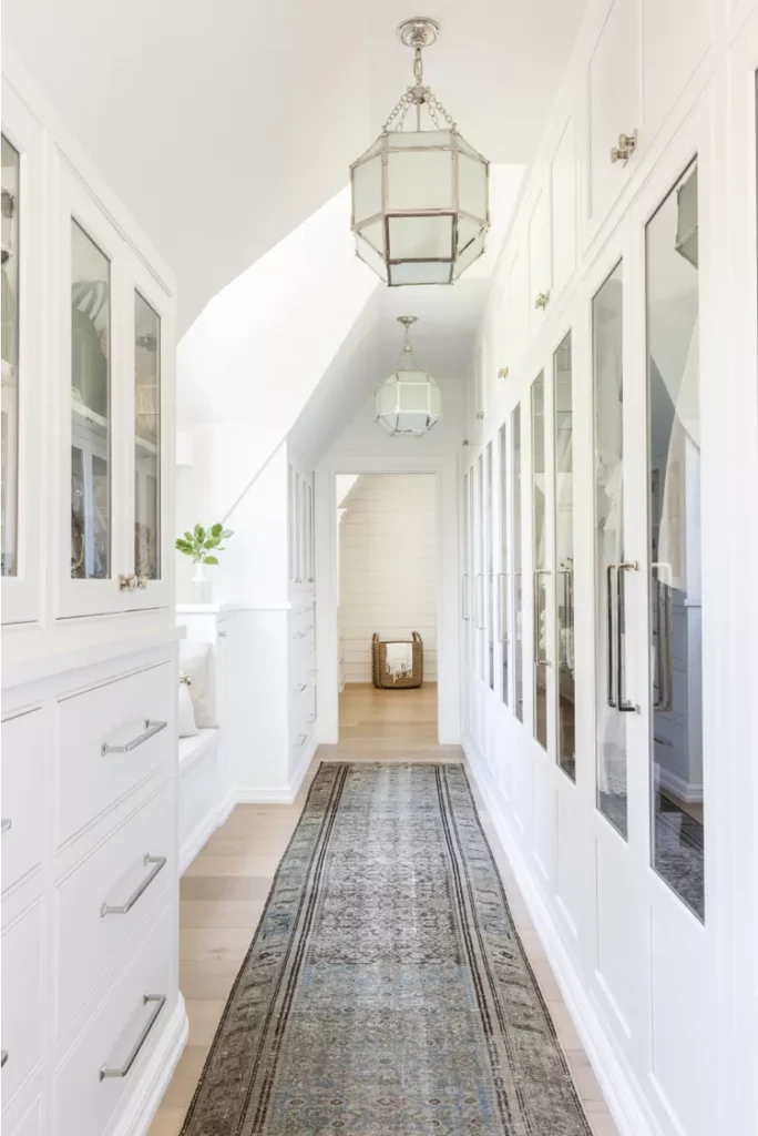A long, bright, and narrow attic hallway transformed into a luxurious walk-through closet, featuring a muted vintage runner rug on light wood flooring. The sloped, pitched roof structures are illuminated by classic lantern pendant lights. The layout maximizes the narrow corridor by lining both sides with seamless custom white built-in dressers, display upper cabinets, and floor-to-ceiling mirrored closet doors to provide low-profile storage that keeps the floor space clear.