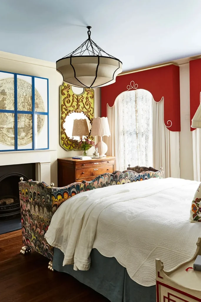 An eclectic luxury bedroom with a uniquely patterned upholstered bed frame, a statement pendant light, and dramatic red shaped pelmets framed with white piping over sheer curtains, demonstrating how To complete your window dressing with a polished, high-end finish, adding a customized valance or upholstered pelmet board provides the perfect crowning touch. Valances and pelmets neatly conceal curtain hardware, tracks, and roller shade mechanisms while adding a sophisticated structured framework to the top of your window. They bring a timeless, tailored architectural feel that makes your bedroom decor look entirely custom-designed. Upholstered pelmets: Box frames wrapped in matching drapery fabric create a clean, modern aesthetic. Conceal functional gear: Perfect for hiding heavy-duty blackout roller blind mechanisms or motorized tracks. Proportional balance: Keep pelmet heights proportional to your window height so they don't block too much natural light.