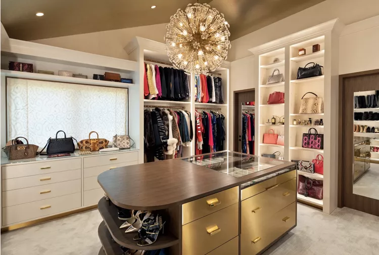 A luxurious, large walk-in closet featuring a sloped ceiling and an ornate gold starburst chandelier over a center island with brass drawers and integrated shoe shelves. Custom white built-in storage units perfectly combine open clothing hanging rails, deep white drawers, and tall display shelving filled with colorful designer handbags, beautifully showcasing diverse attic closet ideas angled ceilings to maximize space under different wall heights.