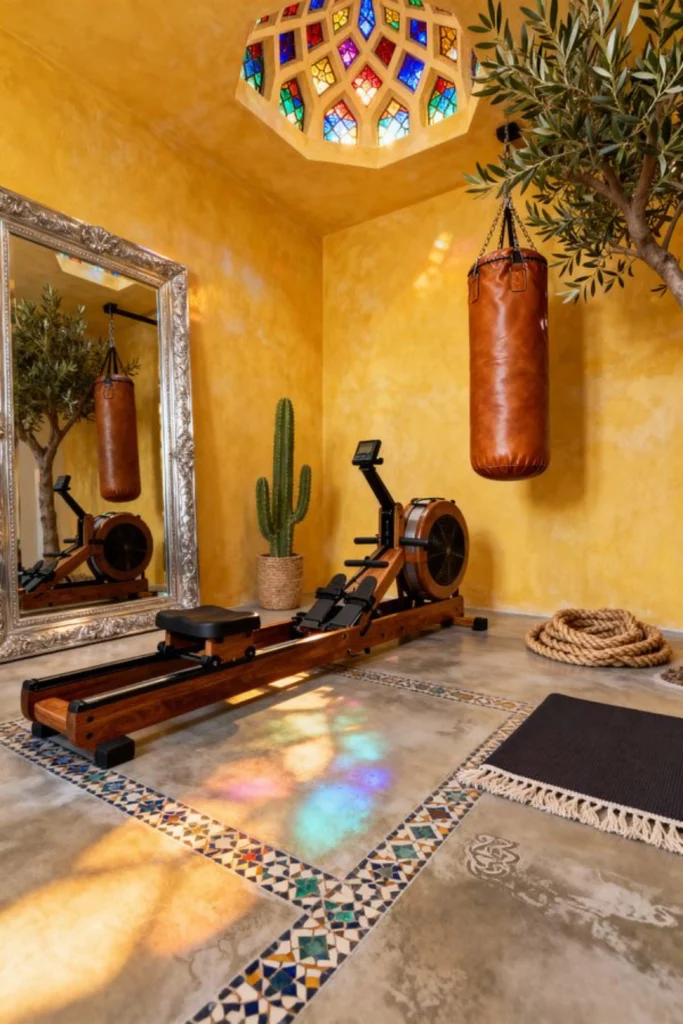 A warm, Mediterranean-style home gym featuring a large ornate floor-standing mirror leaning against yellow plaster walls, reflecting a wooden rowing machine, leather heavy punching bag, stained-glass skylight, and indoor plants.