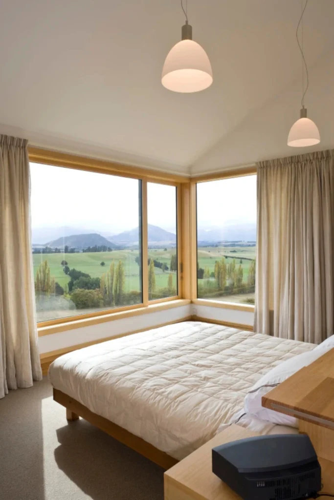 A master bedroom with large corner glass windows overlooking a scenic mountain landscape, featuring full floor-to-ceiling beige linen drapes framing the view, demonstrating how Grand spaces demand grand design choices. If you're lucky enough to have expansive glass or sliding doors, discovering wide window treatment ideas master bedrooms need is all about playing with scale and dramatic proportions. For wide master bedroom windows, single panels will look skimpy and out of place. Instead, install wall-to-wall drapery tracks or extra-wide ceiling-mounted rods. Hanging multiple full-bodied panels allows you to frame the large view beautifully while maintaining a sophisticated, hotel-luxury aesthetic. Ceiling-to-floor drapes: Mount tracks directly to the ceiling so the fabric flows all the way down to the floor. Motorized shades: Combine sheer wide panels with motorized solar shades for effortless daily light control. Rich textures: Velvet, heavy linen, and lined jacquard fabrics elevate large master suites instantly.