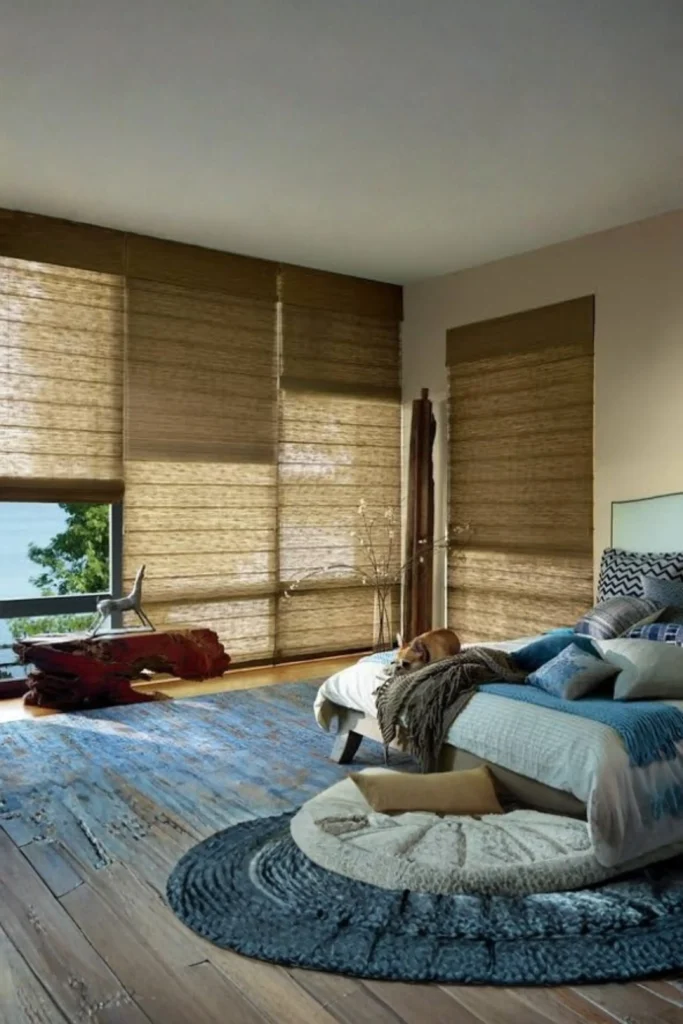 A coastal-style bedroom with a blue patterned area rug and large floor-to-ceiling windows fitted with natural woven bamboo Roman shades filtering warm sunlight, demonstrating how If your bedroom style leans toward Boho, Organic Modern, or Coastal Chic, woven wood and bamboo shades bring rich warmth and earthy texture to your windows. Woven shades look stunning on their own, but pairing them with neutral curtain panels adds a layered, designer touch. They filter sunlight in a warm, dappled pattern while adding an organic grounding element to modern or all-white bedrooms. Opt for privacy liners: Choose top-down, bottom-up wood shades lined with blackout fabric for ultimate privacy. Mix wood tones: Match the bamboo shade tone to your bed frame or nightstand wood tones for a cohesive palette. Pair with linen: Layer creamy, floor-length linen drapes over natural bamboo shades to soften the overall window frame.