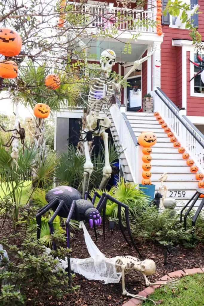 Dramatic Halloween yard setup featuring oversized spiders creeping across your walls and front lawn, with a giant black spider with purple fangs standing over a webbed skeleton, a towering 12-foot skeleton on the porch, orange jack-o'-lantern buckets lining the staircase, and pumpkins hanging from tree branches.