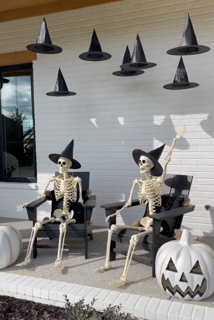 Outdoor Halloween porch decor featuring floating witch hats suspended in mid-air from the porch ceiling above two life-sized skeletons wearing witch hats sitting in black armchairs, paired with white jack-o'-lantern pumpkins on a white brick patio.