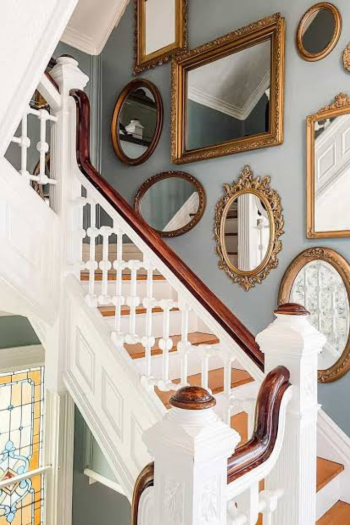 A gallery wall composed of multiple antique gold-framed mirrors in various shapes mounted along a soft grey staircase wall with white wooden railings.