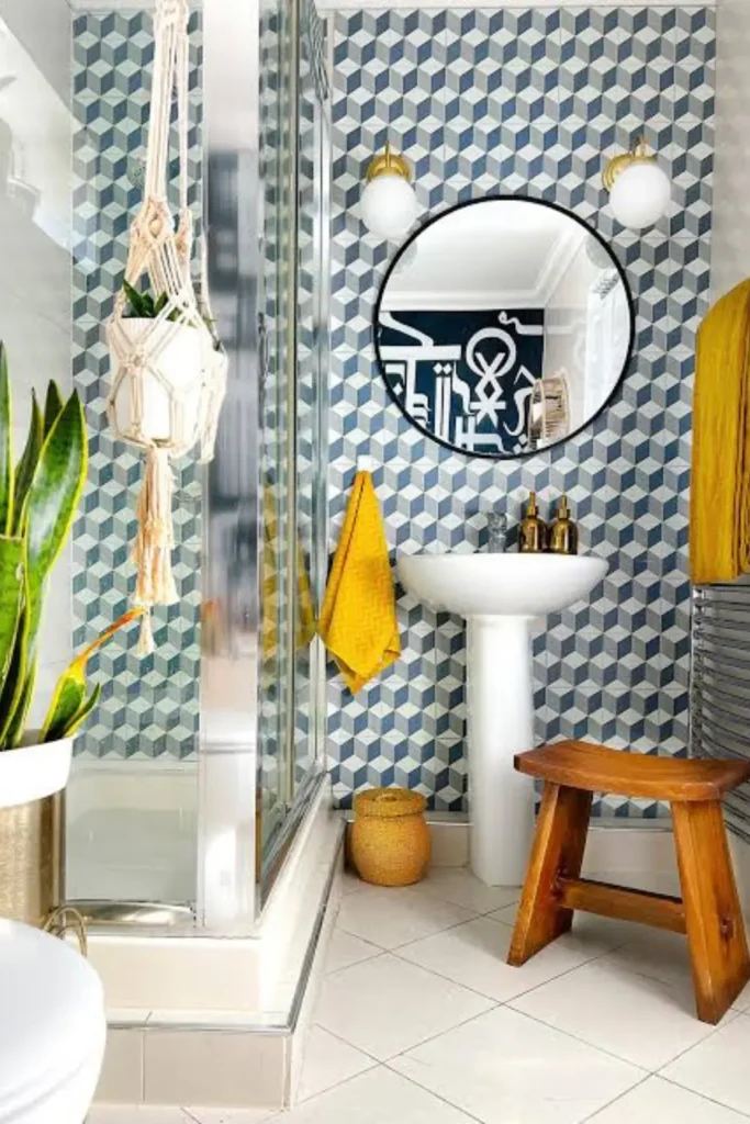 Apartment bathroom ideas on a budget showing geometric blue removable wallpaper, a round black mirror with brass globe wall sconces, a pedestal sink, mustard yellow towels, and hanging macrame plant holders.