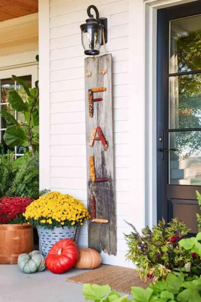 A vertical entrance framing fall porch decor setup featuring a wooden sign spelling FALL with Indian corn cobs hanging under a lantern, paired with yellow mums, red chrysanthemums, and heirloom pumpkins near a black glass door.