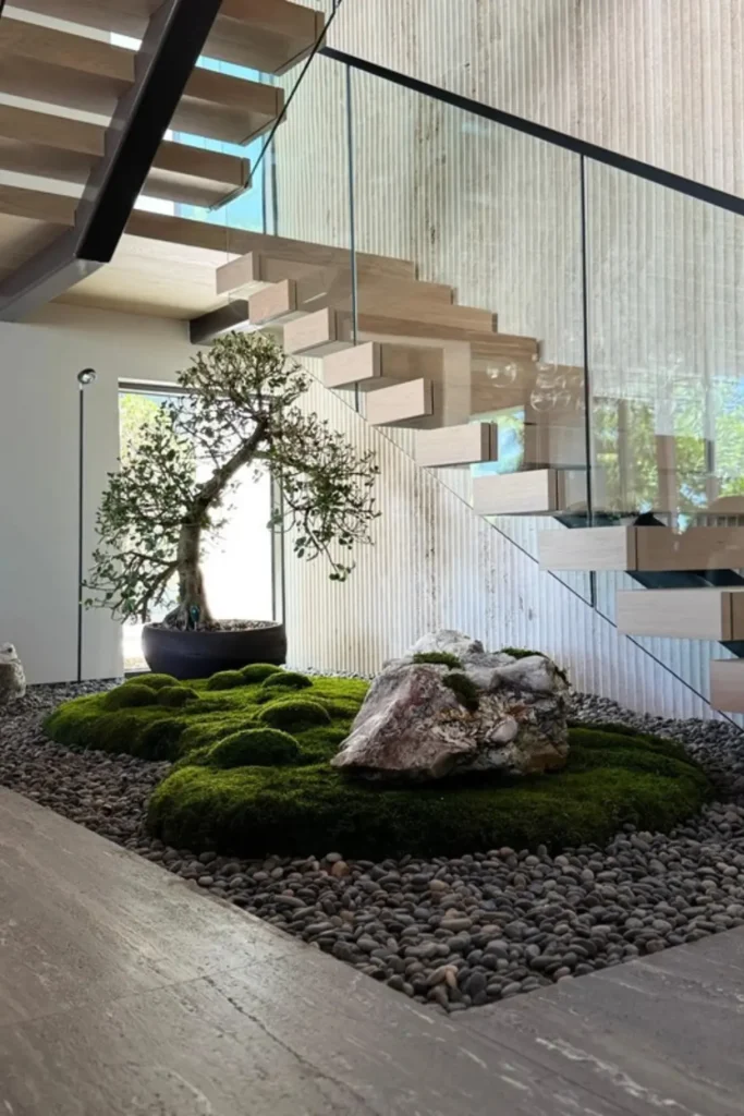 Minimalist Zen rock garden with a dark potted bonsai tree, raised moss mounds, and a large natural stone resting on grey pebbles under modern floating stairs.